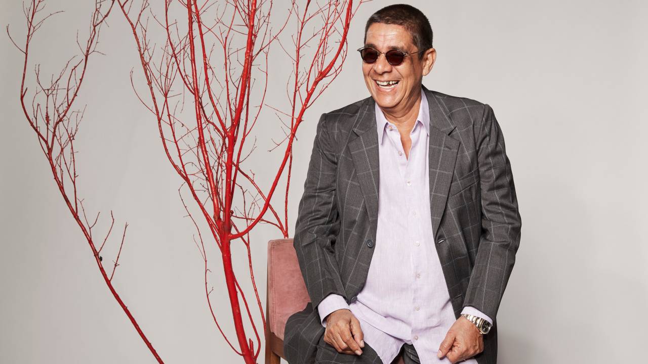Zeca Pagodinho sorri de óculos escuros. Ele está vestindo uma camisa branca e um paletó cinza, sentado numa cadeira