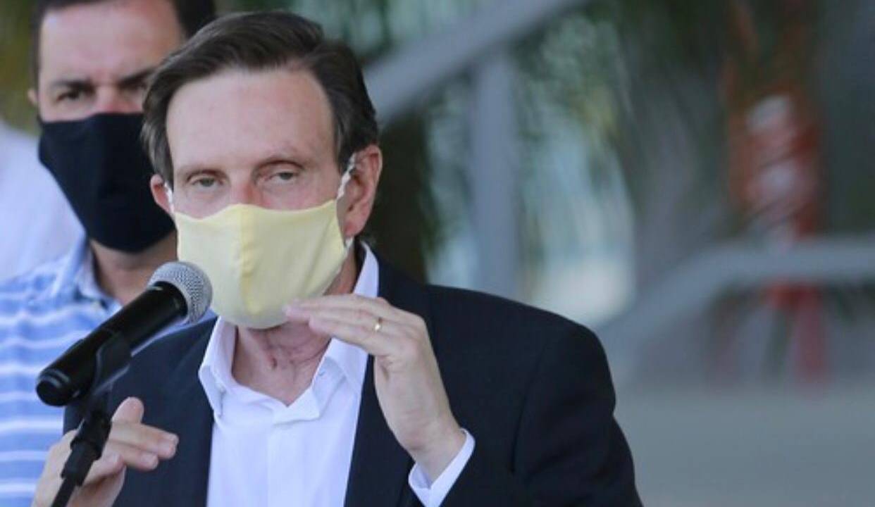 A imagem mostra o prefeito Marcelo Crivella fazendo um discurso, usando máscara de proteção