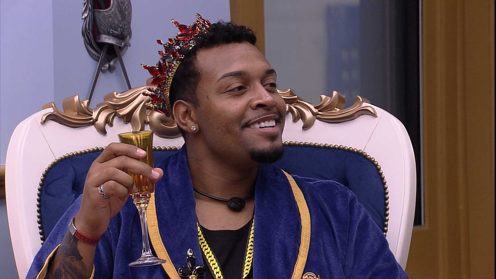 Humorista Nego Di no quarto do líder do BBB 21 com uma coroa na cabeça, roupão azul e segurando taça de champagne