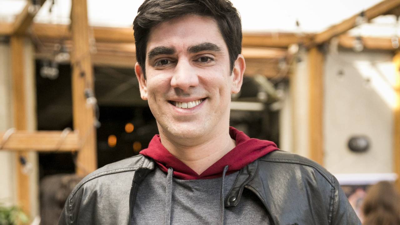 A imagem mostra Marcelo Adnet, de casaco cinza, sorrindo para a câmera