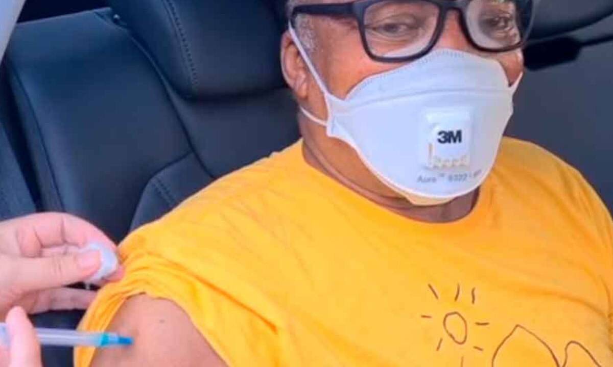 A imagem mostra Milton Nascimento, de óculos, boné, e camiseta amarela, sendo vacinado dentro do carro
