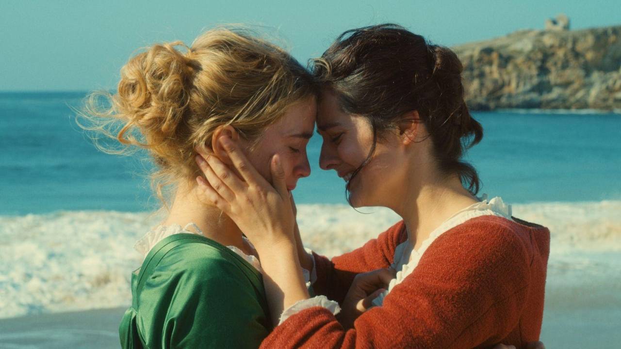 Duas atrizes de testas coladas, roupas de época numa praia