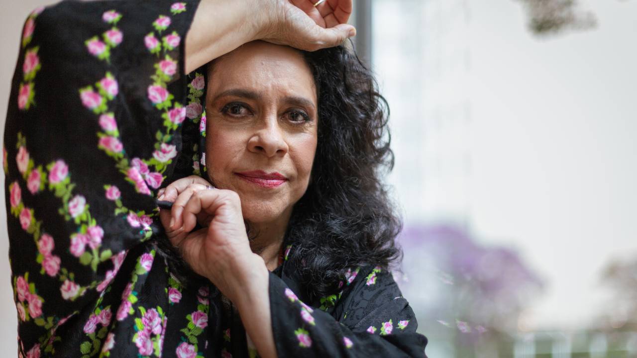 Gal Costa, apoiada numa parede, com uma das mãos acima da cabeça e a outra apoiada no queixo