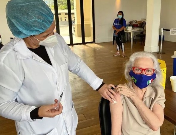 De óculos vermelho e máscara, a atriz Fernanda Montenegro é vacinada por uma agente de saúde usando touca azul