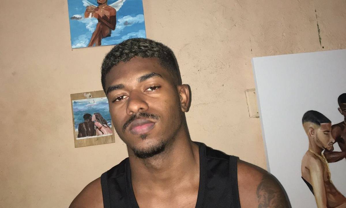 Artista Jota, de regata preta, postando na frente de suas pinturas