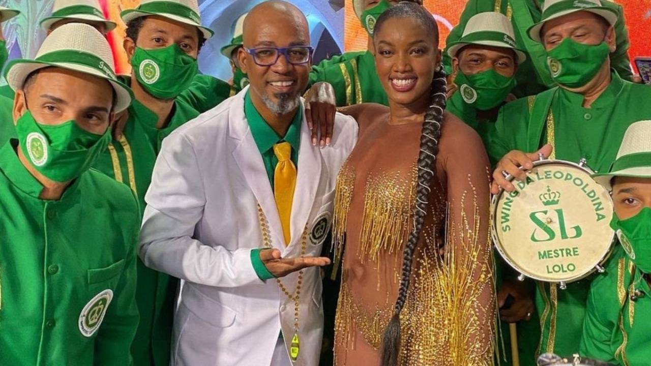 Iza posa sorridente com um macacão dourado de franjas ao lado de músicos da escola Imperatriz Leopoldinense