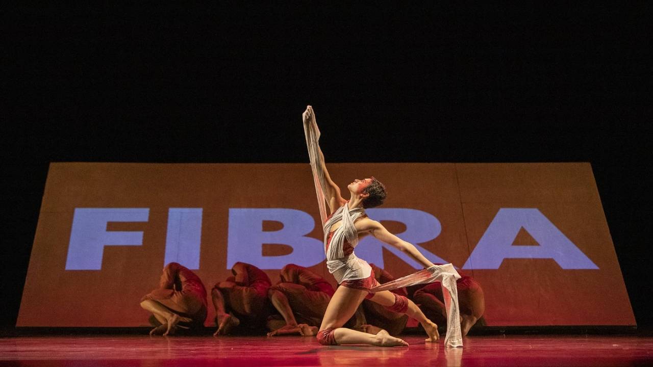 Bailarina da Cia Deborah Colker está no chão em primeiro plano. Em segundo plano, outros cinco bailarinos. Ao fundo, está projetada a palavra Fibra.