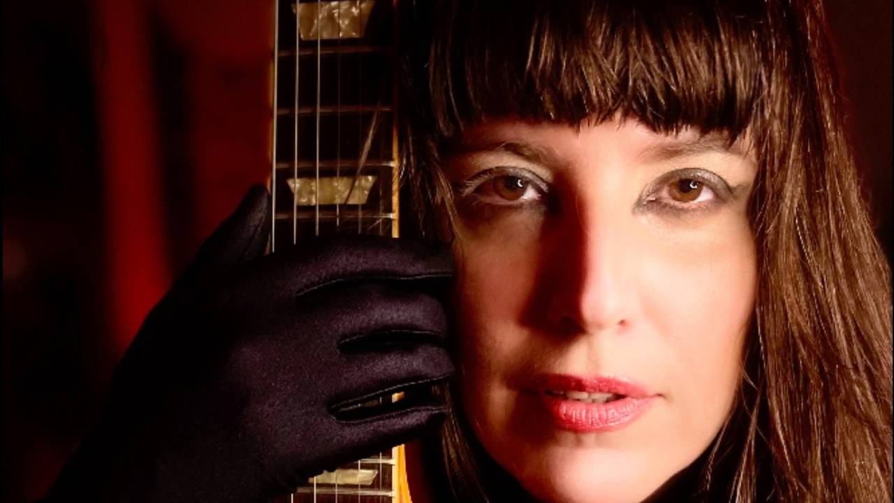A cantora Danni Carlos tem cabelos castanhos com franja. Ela segura o braço de uma guitarra colada a seu rosto, paralelamente, e usa luva preta.