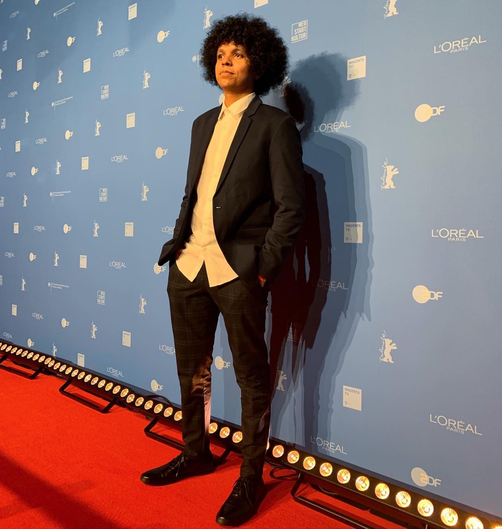 O cineasta Bruno Ribeiro usa cal&ccedil;a cinza xadrez, camisa branca, palet&oacute; cinza e sapato preto. Ele tem cabelo black power e est&aacute; com as m&atilde;os nos bolsos. Est&aacute; sobre o tapete vermelho e ao fundo h&aacute; uma parede azul-claro.