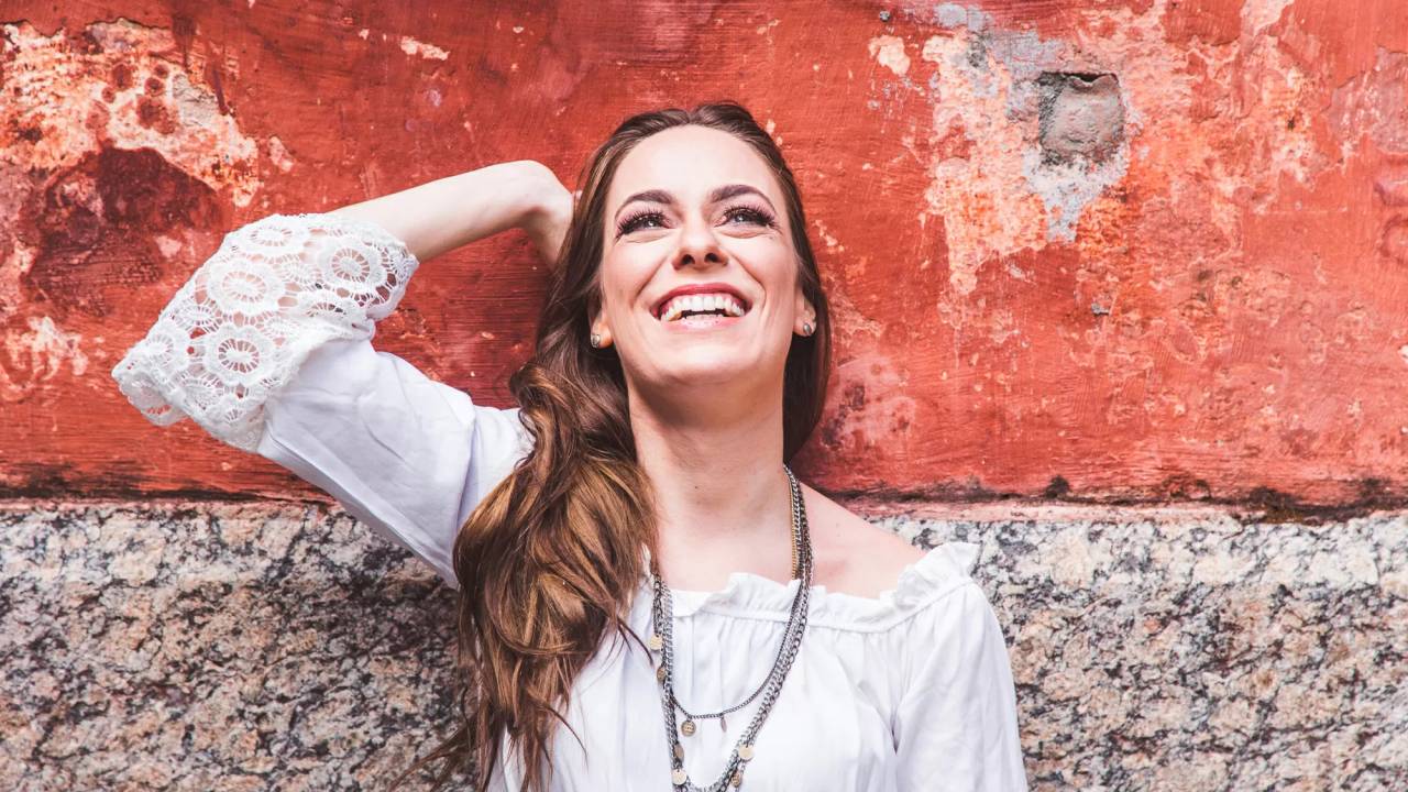 A cantora Laura Zennet é branca e tem cabelos compridos castanhos claros. Ela está de camisa branca bordada e está encostada em uma parede de pedra na parte de baixo e avermelhada na parte de cima. Ela está olhando para o alto e sorrindo. Sua mão direita está encostada na parede, na altura da sua cabeça.