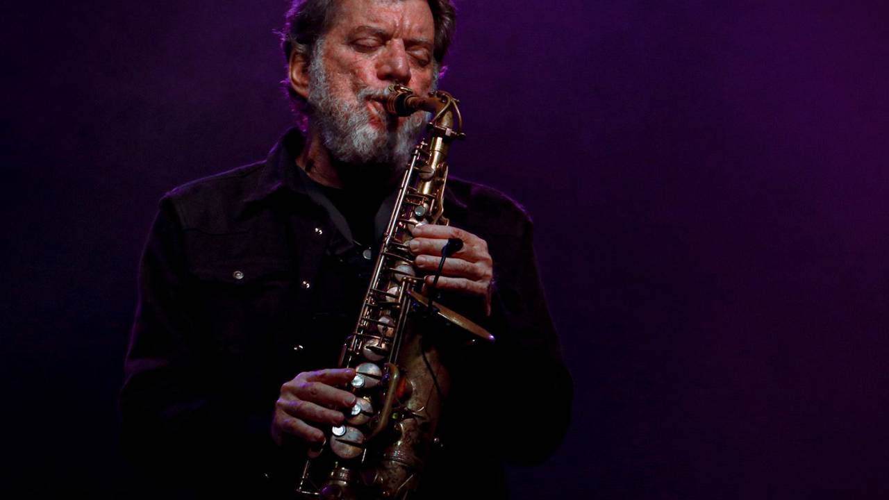 Leo Gandelman está de olhos fechados tocando saxofone. Ele veste jaqueta preta. Ao fundo, uma luz roxa.