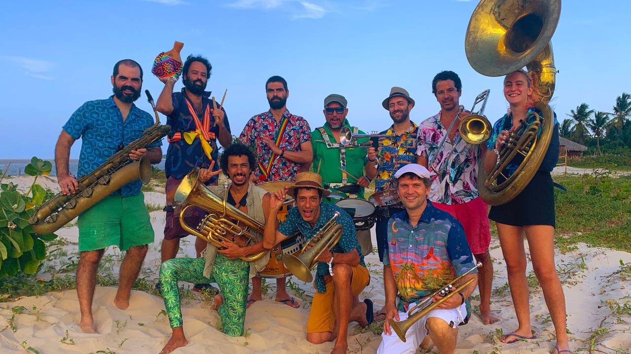 Três integrantes da Orquestra Voadora estão na frente, com seus instrumentos na mão, com um dos joelhos apoiados no chão. Atrás, estão sete, em pé, também com seus instrumentos. A única mulher está à direita da foto, em pé, segurando seu sousafone. Todos os músicos estão sorrindo.