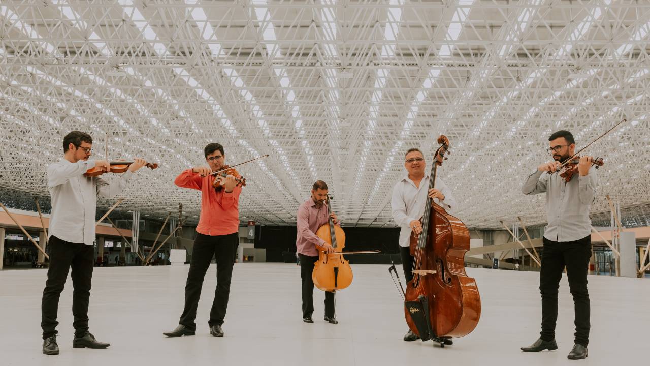 Os cinco integrantes do Quinteto da Paraíba em lugar com chão e teto branco. Da direita pra esquerda: Ronedilk Dantas (1º violino), Ulisses Silva (viola), Nilson Galvão (violoncelo), Xisto Medeiros (contrabaixo) e Anderson Carvalho (2º violino).