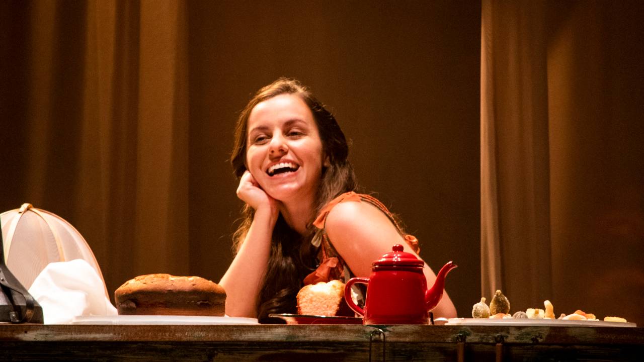 Raquel Penner é branca e tem cabelos castanhos lisos. Ela está sorrindo, com a mão direita apoiada no rosto e o cotovelo na mesa, um móvel de madeira que tem um bule vermelho, bolo e frutas cristalizadas sobre ele.