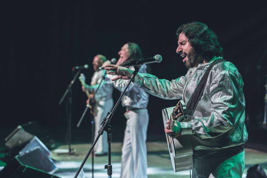 Geminis Bee Gees 07 – Divulgação Eduardo Alexandre-min Três integrantes do grupo Geminis Bee Gees com roupas prateadas, cantando em frente aos respectivos microfones.