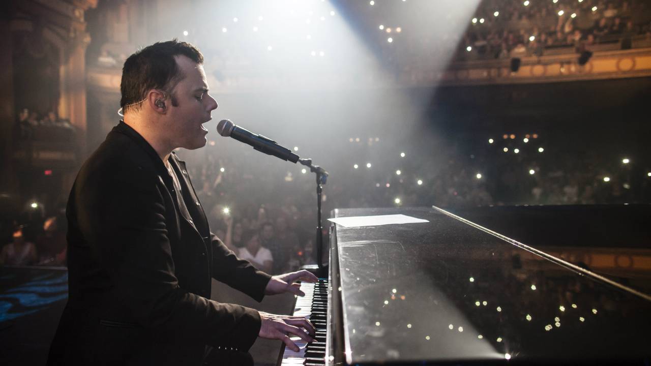 O canadense Marc Martel veste roupa preta e está sentado, cantando e tocando um piano de cauda. Ao fundo, as luzes da plateia.