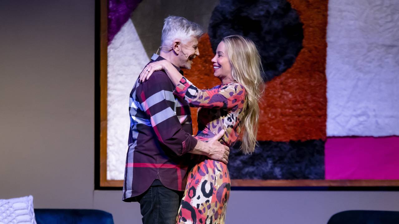 Miguel Falabella e Danielle Winits estão no palco, se olhando e sorrindo. Ele está com a mão na cintura dela. Ela tem a mão apoiada no ombro deles.