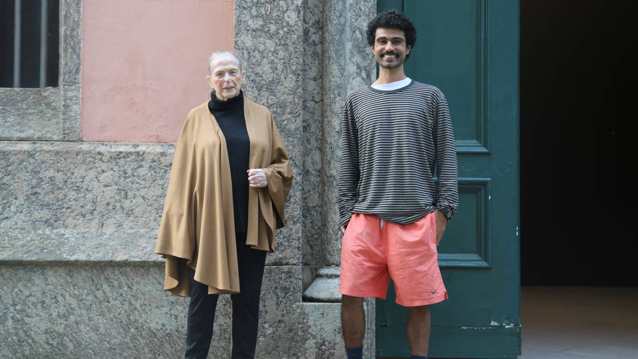 Katie van Scherpenberg de calça e blusa pretas e casaco bege. Bernardo Magina de bermuda rosa e casaco cinza.