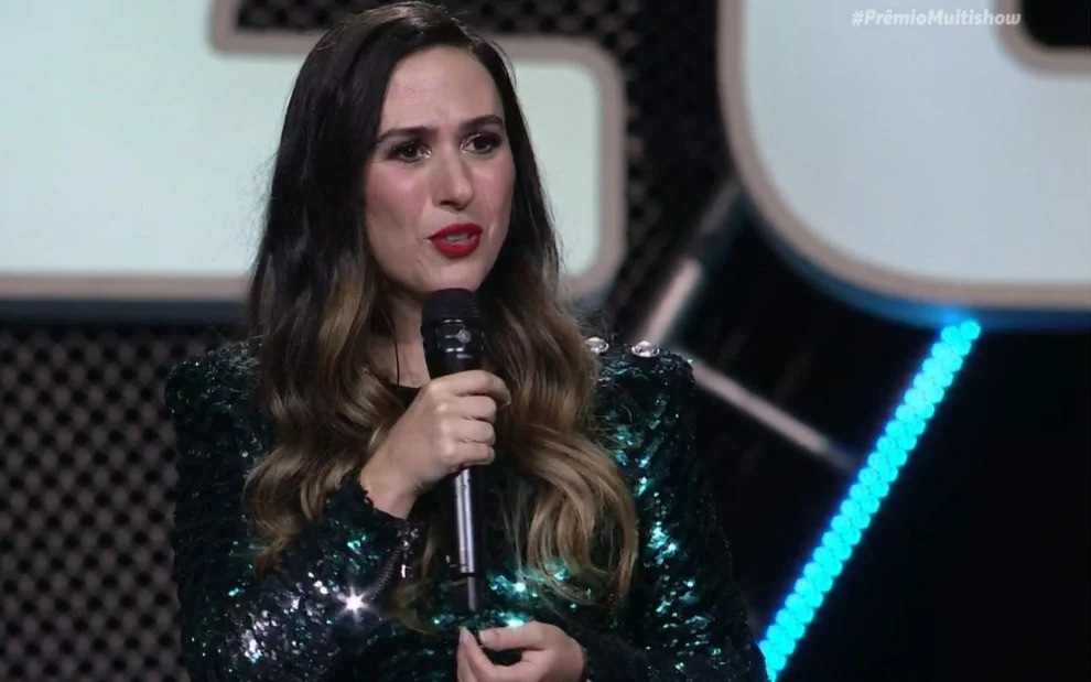 Tatá Werneck de vestido de paetê preto com detalhes verdes, com microfone na mão, apresentando o Prêmio Multishow 2020.