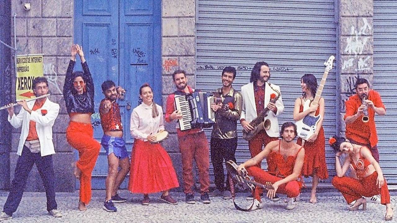 Os onze músicos, com peças de roupa vermelhas na frente de um sobrado antigo