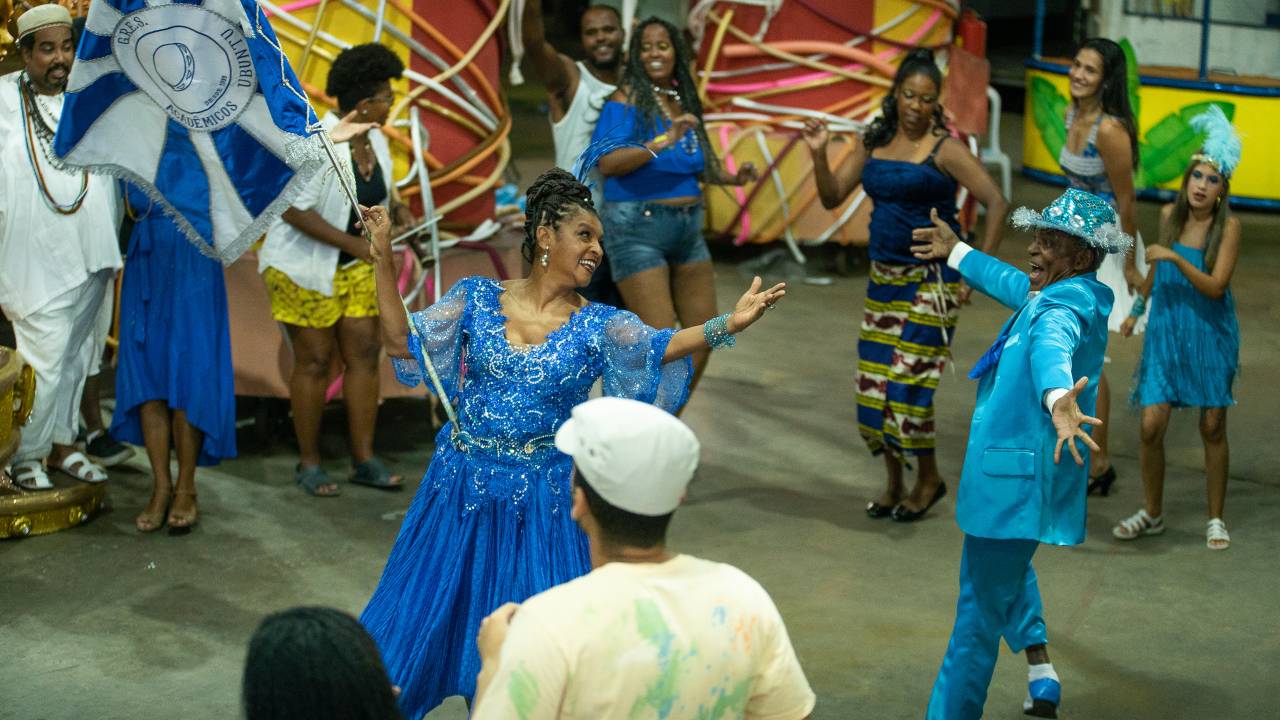 Elisa Lucinda é a porta-bandeira e está à esquerda, segurando o pavilhão de sua escola de samba. Ela usa vestido azul. Antonio Pitanga, à direita, é o mestre-sala, e usa terno azul um pouco mais claro.