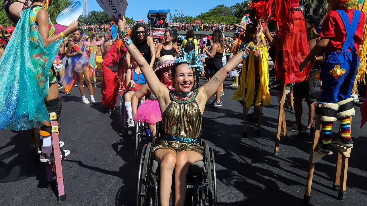 Mulher está sentada em cadeira de rodas e com os braços abertos para o alto, cercada por pernaltas na Orquestra Voadora. Atrás dela, é possível ver outras duas pessoas cadeirantes, em fila.