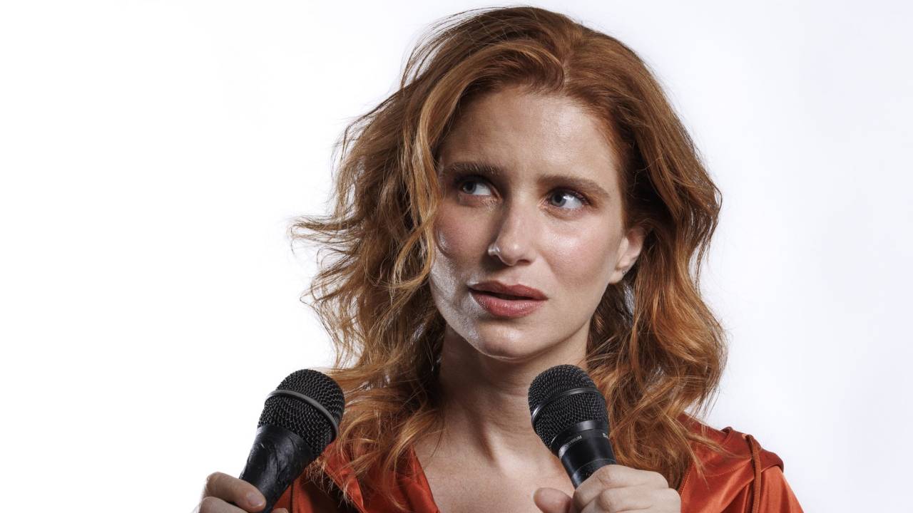 Julia Lund é ruiva, segura dois microfones e veste um moletom marrom.