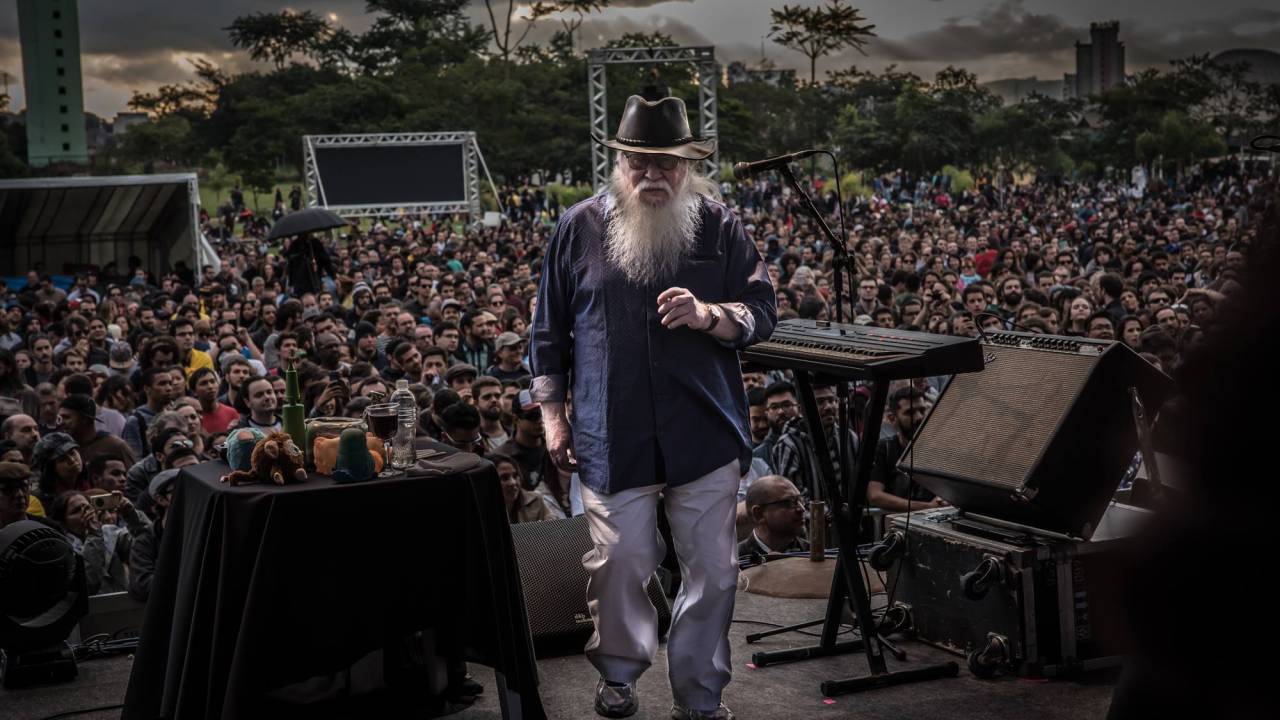 Hermeto Paschoal é albino e tem barba e cabelos compridos e brancos. Ele usa chapéu e está no palco, de costas para o público.