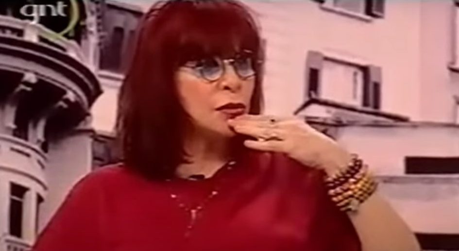 Rita Lee usa camisa em tom de vermelho escuro e tem a mão apoiada abaixo da boca