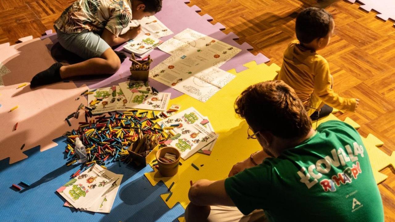 Foto mostra crianças pintando desenhos com giz de cera acompanhadas de um instrutor.