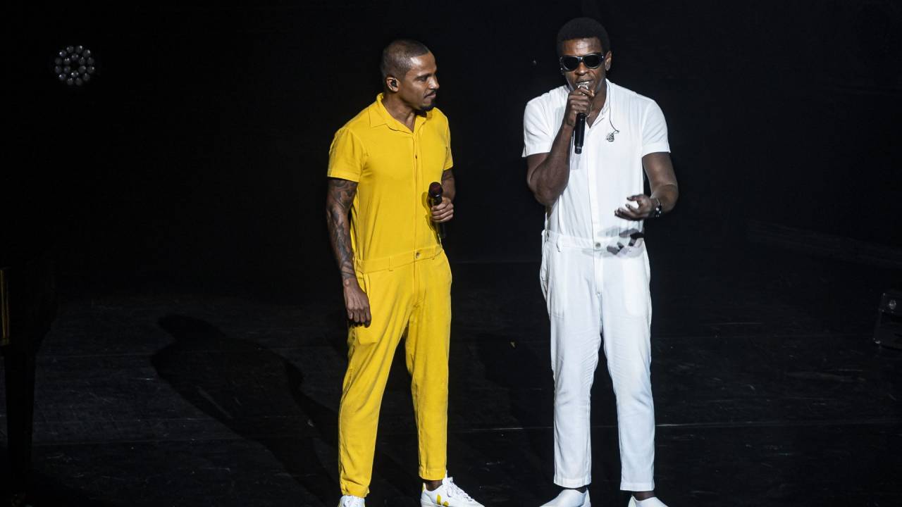 Alexandre Pires é um homem negro de cabelo raspado, ele está de pé, veste macacão amarelo, tênis branco, segura um microfone e olha para Seu Jorge, homem negro de cabelo curto, que usa óculos escuros, macacão e tênis brancos e está cantando.