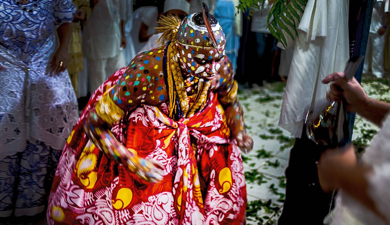 Imagem de um iaô (filho de santo) da orixá Ewá, uma pessoa negra vestida com roupa ritual vermelha, com cabeça raspada e pintura de pontos colorido pela cabeça, colo e braços