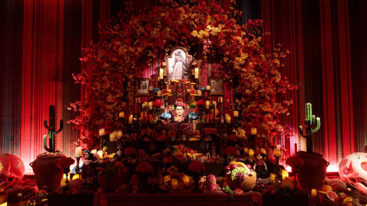 Com um fundo vermelho, imagem traz estante com diversas imagens de Frida Kahlo, frutas e velas