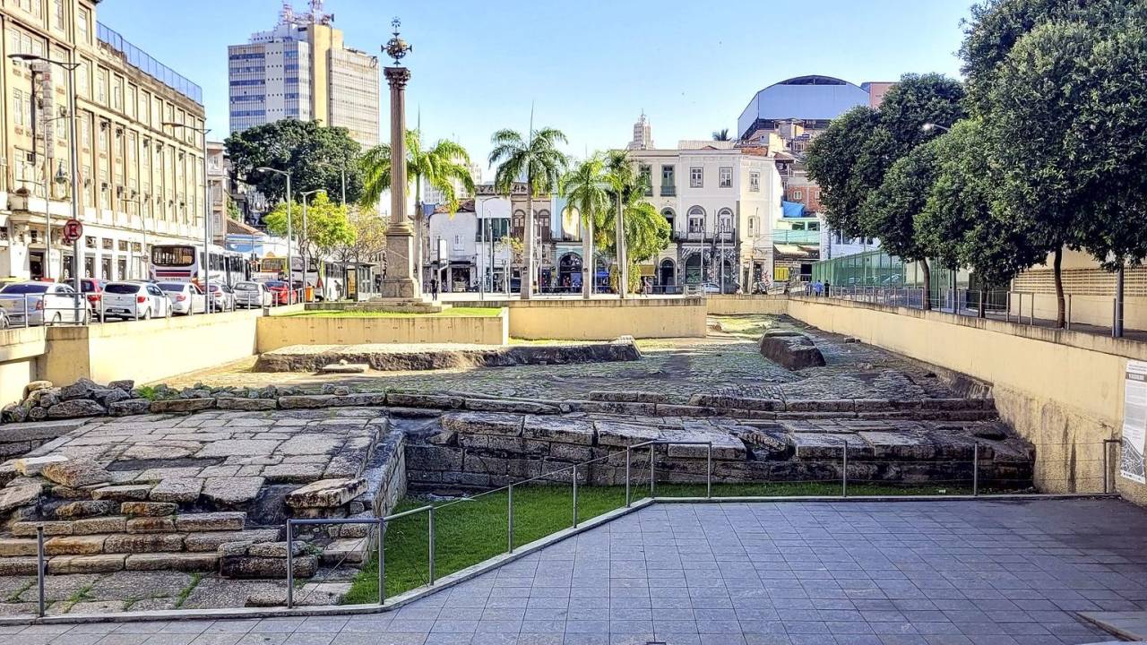 Imagens do Cais do Valongo, em pedra, com o casario histórico da Gamboa ao fundo e o céu azul.