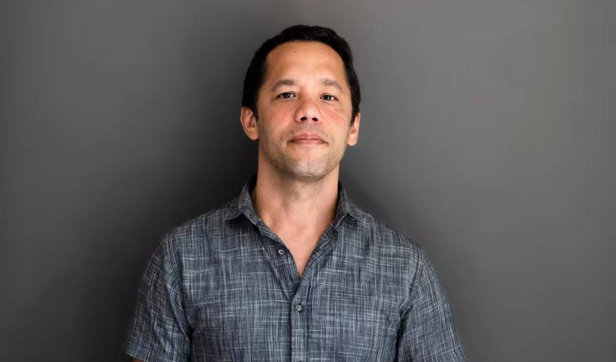 Itamar Vieira Junior é um homem negro de pele clara, de cabelos ondulados e curtos. Ele usa camisa de botão de mangas curtas cinza, e está escorado em uma parede cinza.