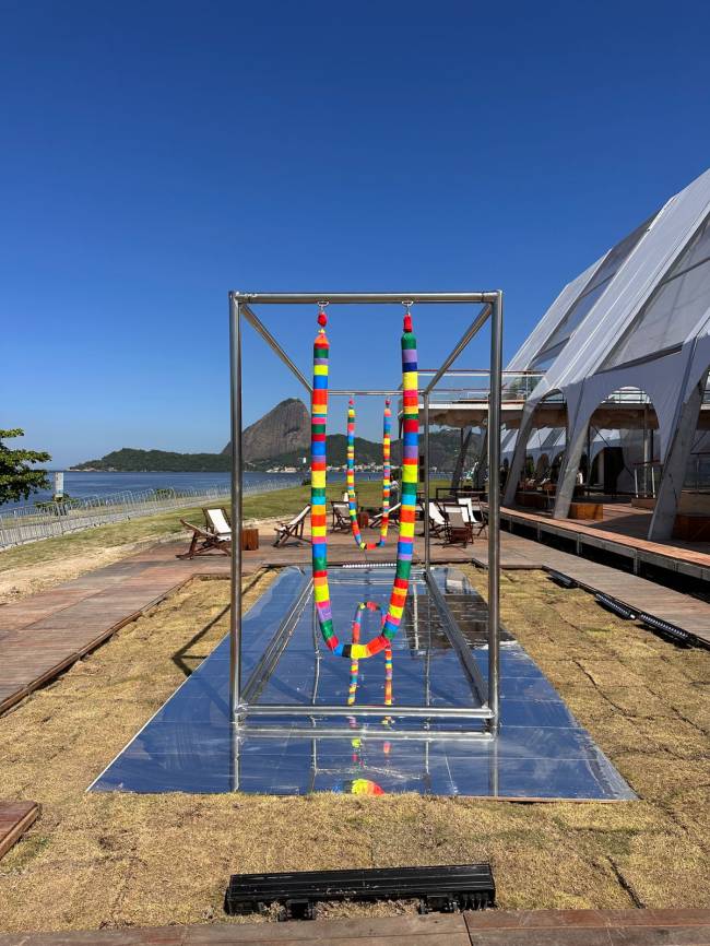 Balanço em aço com cordas revestidas de tecidos coloridos. Ao fundo, de longe, o céu azul e o Morro da Urca.