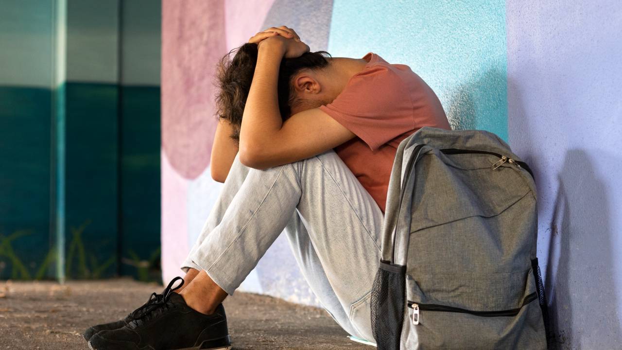 Jovem leva as mãos a cabeça escondendo o rosto sentado no chão, ao lado de uma mochila.