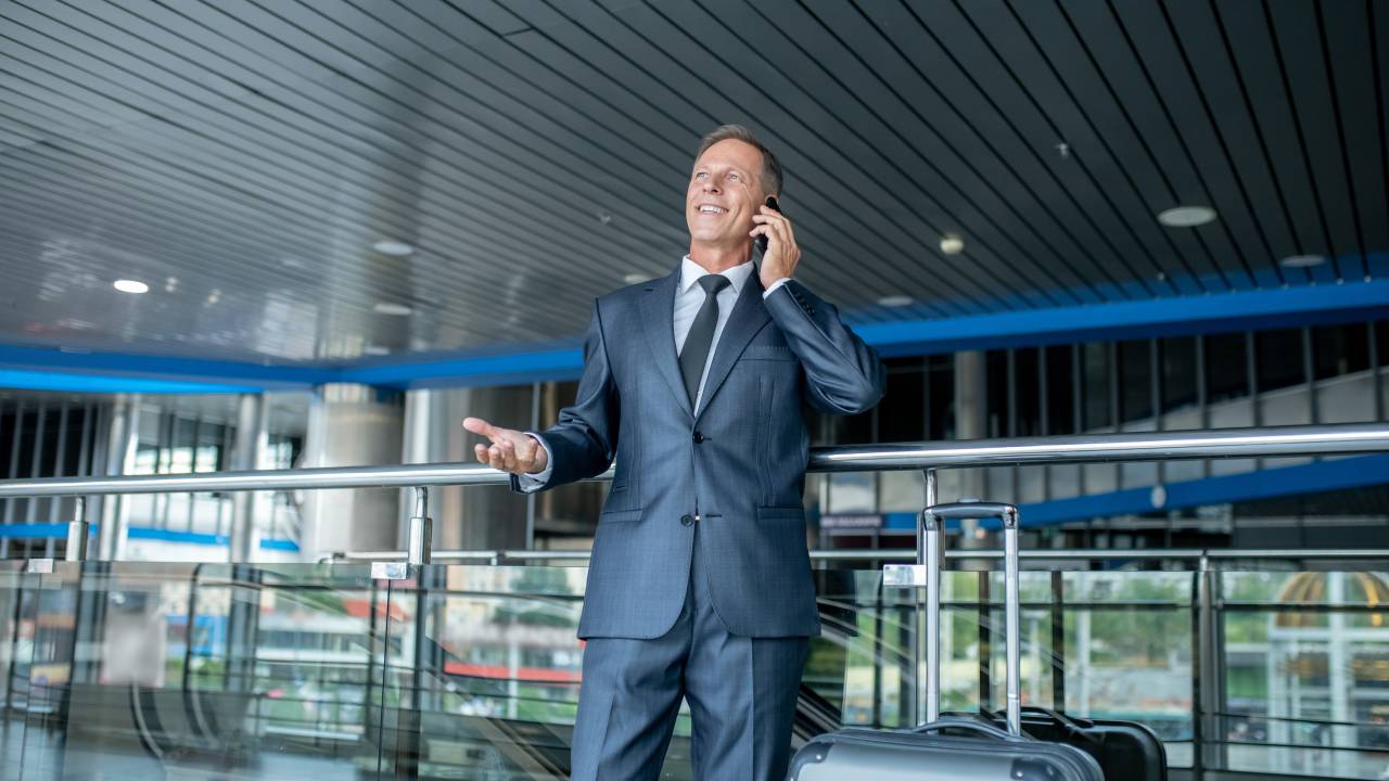 Homem de pé no aeroporto ao telefone e ao lado de uma mala de viagem.