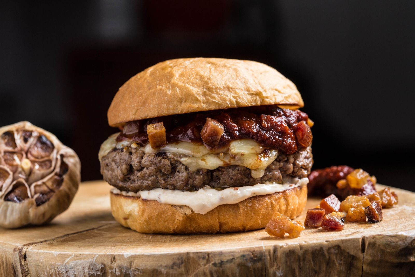 Hamb&uacute;rguer gourmet com p&atilde;o brioche, carne, queijo derretido, molho vermelho e bacon em cubos, sobre uma t&aacute;bua de madeira r&uacute;stica, com uma cabe&ccedil;a de alho assado &agrave; esquerda
