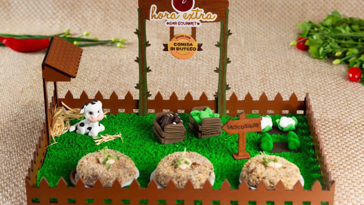 Petisco de costela do Hora Extra, servido em uma Miniatura de fazenda com cerca de madeira, gramado verde, uma vaquinha, feno, caixotes com salgados, e uma placa "Hora Extra Bar Gourmet Comida de Buteco"