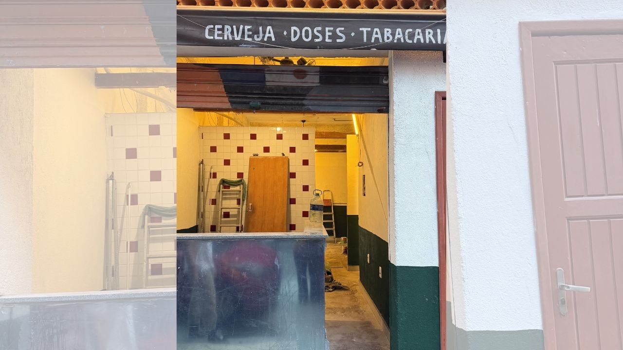 Fachada de um bar ou tabacaria em reforma, com letreiro "CERVEJA DOSES TABACARIA". O interior tem paredes brancas com azulejos vermelhos e brancos, uma porta de madeira, escadas e um balcão metálico. À direita, uma porta rosa clara.