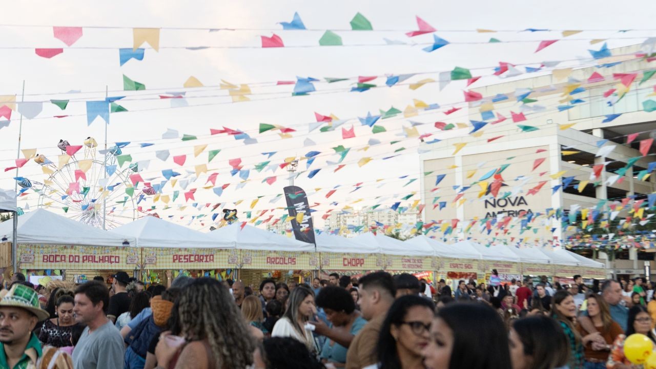 Multidão em festa junina com barracas de "Boca do Palhaço", "Pescaria", "Arraiá" e "Chopp", sob bandeirinhas coloridas e roda-gigante ao fundo, com o letreiro "Nova América" em um prédio