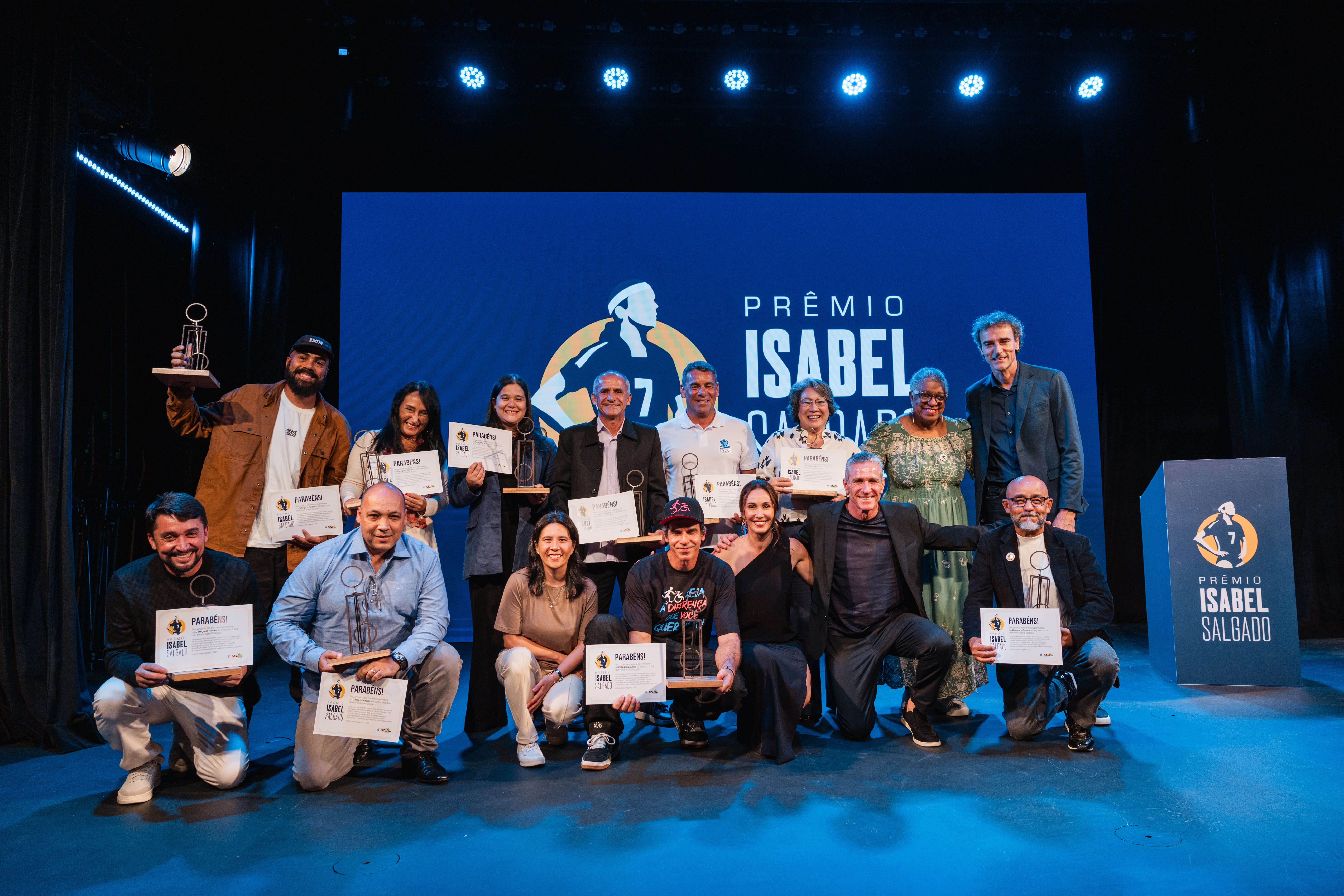 Um grupo de 15 pessoas, homens e mulheres de diversas idades, posa sorridente no palco de um evento. A maioria segura certificados e trof&eacute;us, celebrando o 