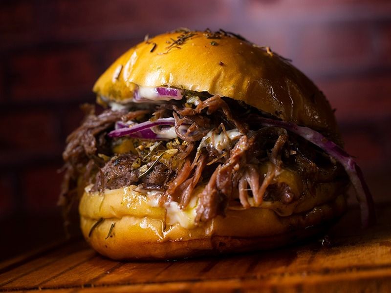 Hamb&uacute;rguer gourmet com p&atilde;o brioche dourado, carne desfiada, queijo derretido, cebola roxa e molho branco, sobre t&aacute;bua de madeira com fundo de tijolos escuros