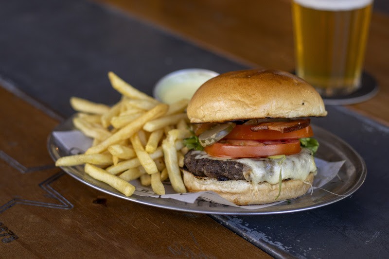 Hamb&uacute;rguer com p&atilde;o brioche, carne, queijo derretido, tomate, alface e picles, acompanhado de batatas fritas e um copo de cerveja, sobre uma mesa de madeira e metal