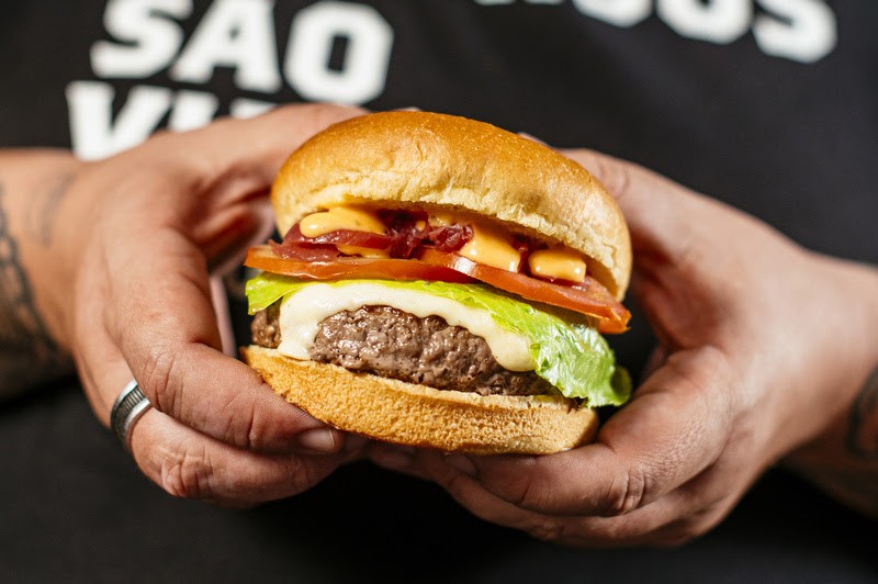 M&atilde;os segurando um hamb&uacute;rguer com p&atilde;o brioche, carne, queijo derretido, alface, tomate e molho especial. O fundo &eacute; escuro com letras brancas desfocadas