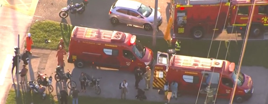 Vista a&eacute;rea de uma rua com viaturas vermelhas de bombeiros e ambul&acirc;ncias, al&eacute;m de um carro prata e v&aacute;rias motos. Pessoas, algumas vestindo uniformes de bombeiro, est&atilde;o pr&oacute;ximas aos ve&iacute;culos, indicando uma opera&ccedil;&atilde;o de emerg&ecirc;ncia ou resgate. O cen&aacute;rio &eacute; urbano, com cal&ccedil;adas e vegeta&ccedil;&atilde;o lateral