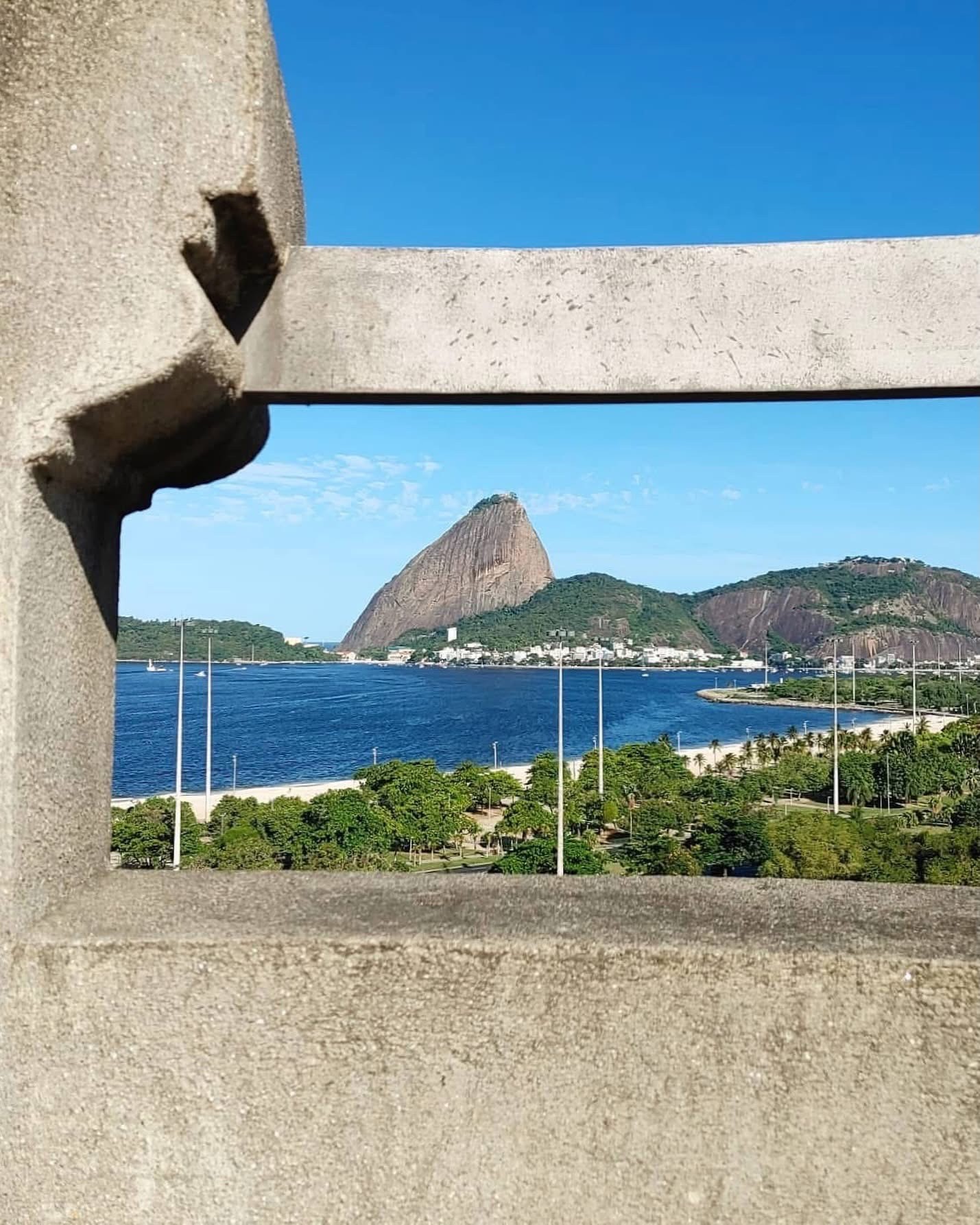 Vista do Pão de Açúcar e da Baía de Guanabara no Rio de Janeiro, emoldurada por uma estrutura de concreto. O céu azul claro contrasta com o mar escuro, e a orla tem vegetação verde e postes de luz. Pequenas construções aparecem na base da montanha.