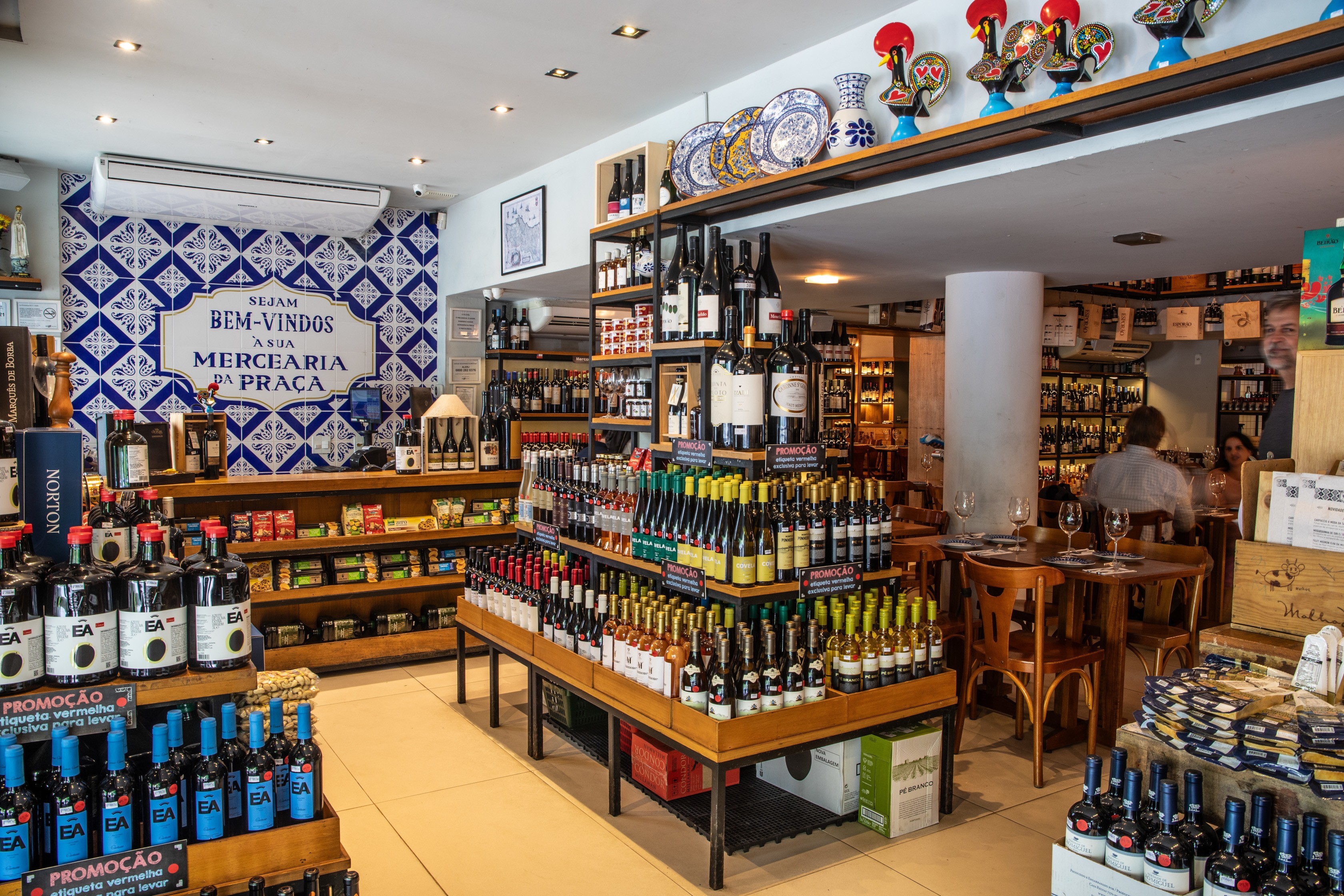 Interior de uma mercearia com prateleiras repletas de garrafas de vinho e produtos gourmet. Ao fundo, uma parede decorada com azulejos azuis e brancos exibe a frase Sejam Bem-Vindos &agrave; Sua Mercearia da Pra&ccedil;a. No lado direito, mesas de madeira com clientes sentados, e um homem de barba grisalha observa a cena. O ambiente &eacute; bem iluminado e acolhedor