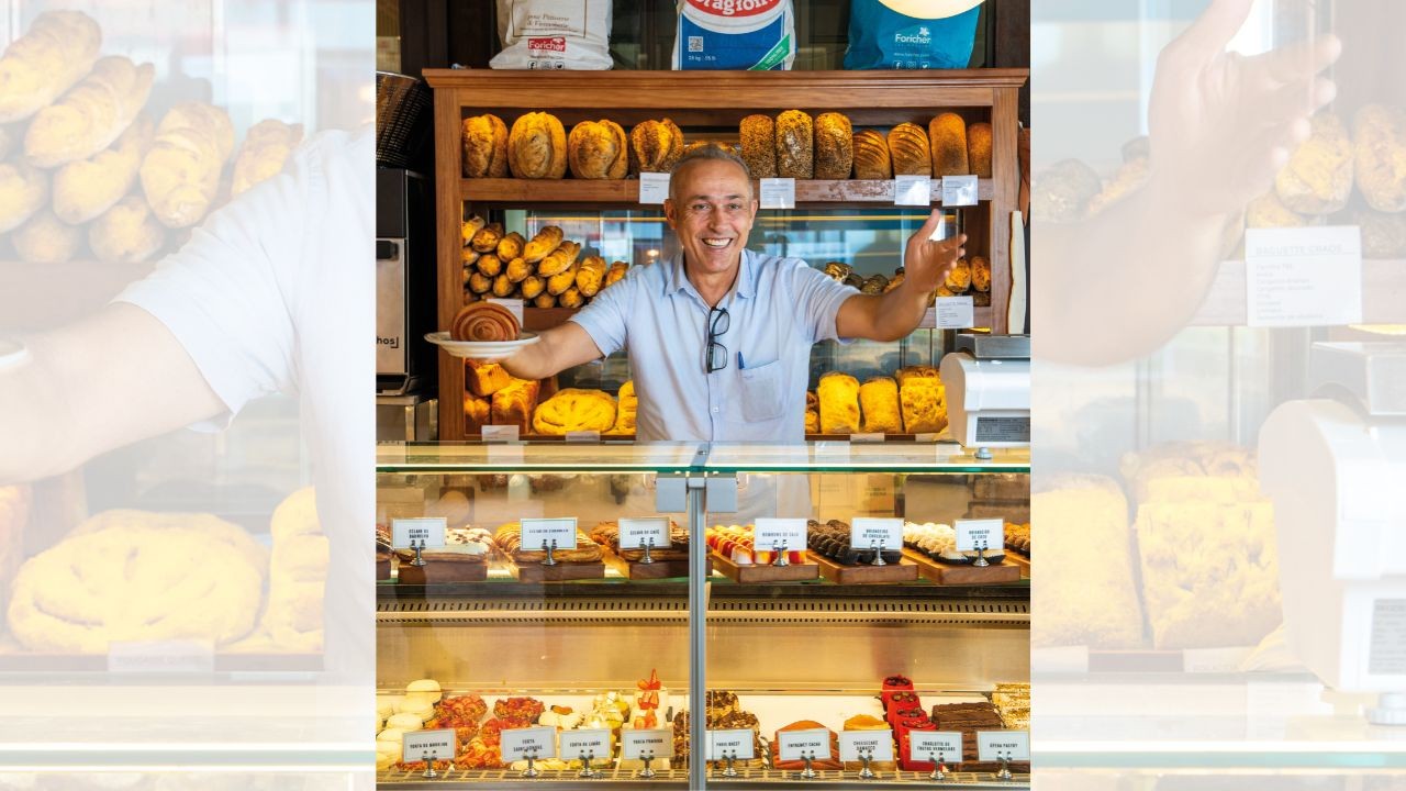 Homem sorridente de camisa azul clara e óculos pendurados, segurando um pão doce em um prato branco, atrás de um balcão de padaria com doces e pães expostos